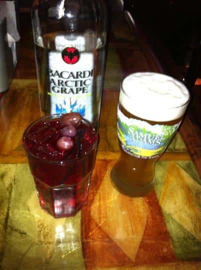 A glass of beer and a glass of a cocktail drink on the bar counter with a Bacardi bottle in the background