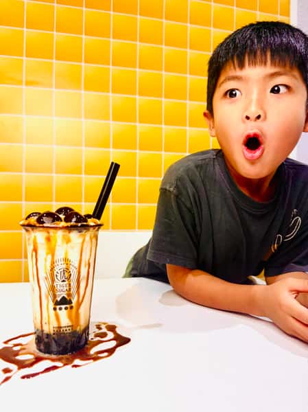 Young boy with a surprised expression sits beside a Tiger Sugar bubble tea with brown sugar syrup dripping on the table.