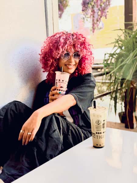 Woman with curly pink hair and round sunglasses smiles while holding bubble tea, sitting casually near a window with plants in view.