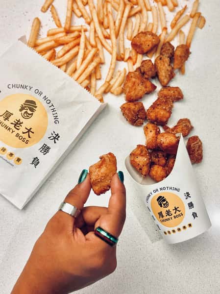 Hand holding crispy fried chicken nugget above Chunky Boss packaging, with French fries and more nuggets scattered on a white surface.