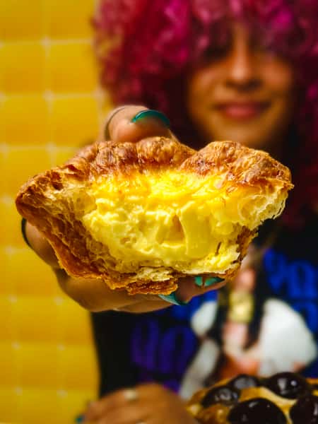 Close-up of a hand holding a flaky egg tart with a creamy yellow filling, person with curly pink hair smiling in the background.