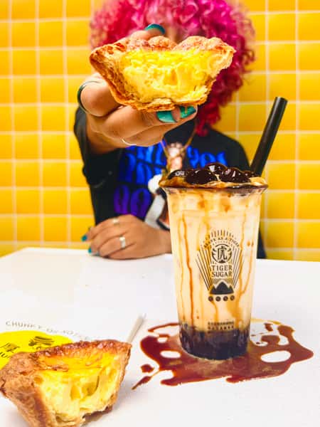Person with pink curly hair holds up a flaky egg tart next to a Tiger Sugar brown sugar bubble tea on a table with yellow tiles.