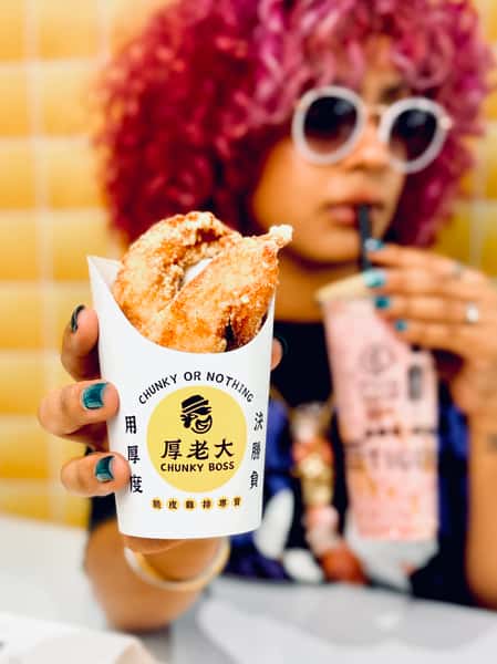 A person with curly pink hair and sunglasses holds crispy fried chicken in Chunky Boss packaging, sipping bubble tea in the background.