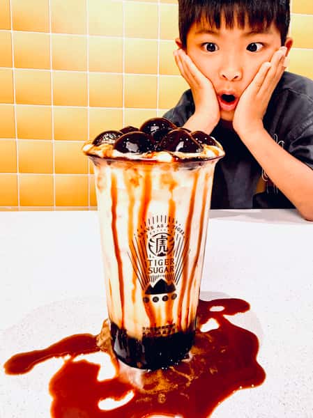 A surprised child stares at an overflowing Tiger Sugar brown sugar bubble tea with tapioca pearls and syrup spilled on the table.