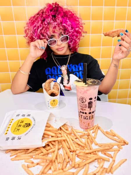Person with curly pink hair and sunglasses holds a piece of fried chicken next to fries, Tiger Sugar bubble tea and Chunky Boss chicken on a table.