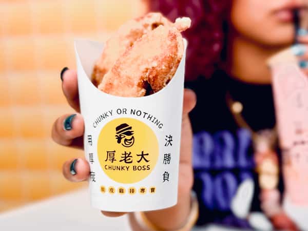 A person holds a serving of crispy fried chicken in Chunky Boss packaging while sipping bubble tea in the background.