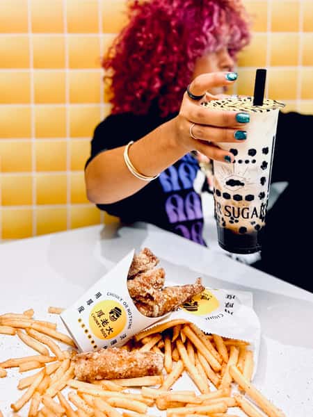 Person with curly pink hair holds a Tiger Sugar bubble tea above a meal of Chunky Boss fried chicken and fries on a white table.