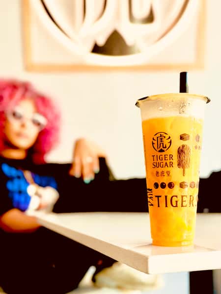 Close-up of a Tiger Sugar mango bubble tea on a table, with a person in the background wearing white sunglasses and pink curly hair.