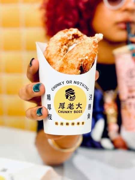 Person with curly pink hair and sunglasses holds up Chunky Boss fried chicken while sipping bubble tea in front of a yellow tiled wall.