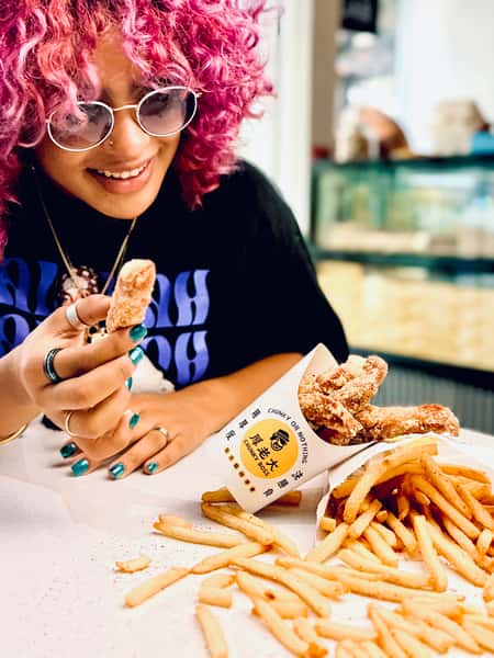 Smiling person with pink curly hair holding a cinnamon churro sits at a table with seasoned fries and a Chunky Boss churro cup.