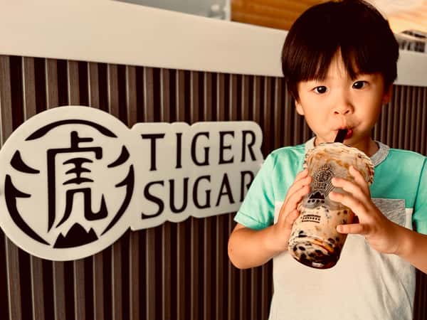 Young boy drinking bubble tea in front of a Tiger Sugar sign with a striped black and white wall background.