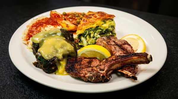 A Greek platter with grilled lamb chops, lemon slices, spanakopita, dolmades with avgolemono sauce, rice, and roasted potatoes.