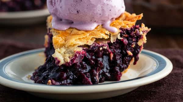 Slice of blueberry pie with a flaky crust, topped with melting purple ice cream, on a white plate with a blue rim.