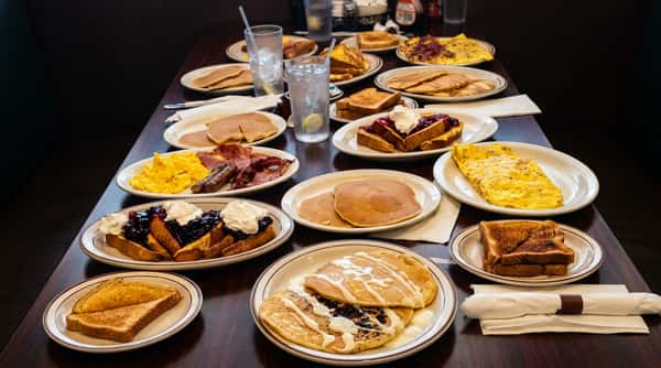 A table overflowing with various breakfast dishes, including pancakes, scrambled eggs, ham, sausage, toast, and French toast.