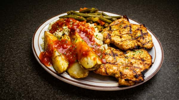 Two grilled chicken breasts with rice, roasted potato wedges, green beans, carrots, and a red sauce, served on a white plate with a brown rim.