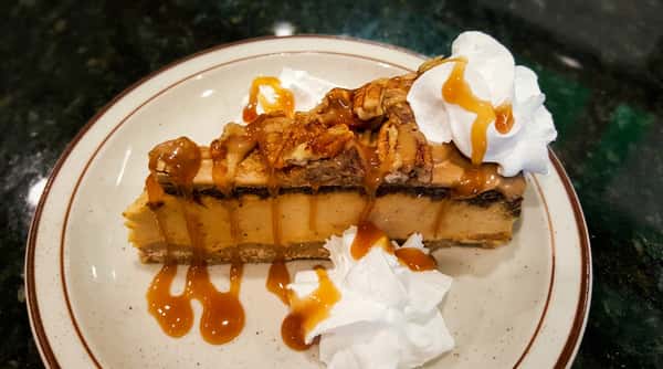 Slice of pecan caramel cheesecake with whipped cream and caramel drizzle on a white plate.