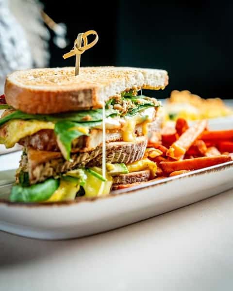 Gourmet grilled sandwich stacked with melted cheese, fresh greens, and sliced vegetables on toasted multigrain bread, served with crispy sweet potato fries on a modern restaurant plate with a warm, casual dining ambiance.