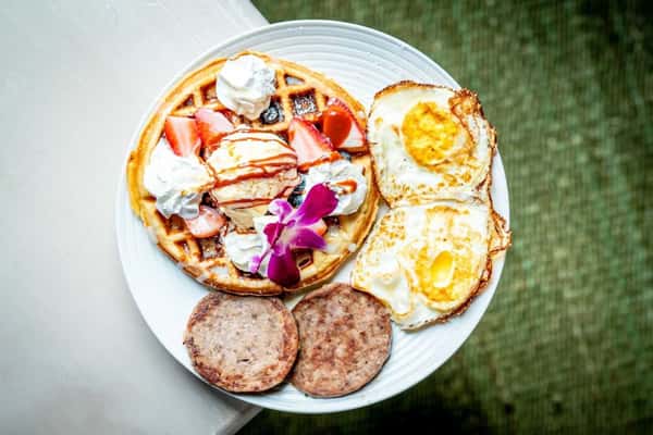 Classic waffle breakfast topped with fresh strawberries, whipped cream, and syrup, served with eggs and sausage for a comforting diner-style meal.