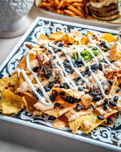 Loaded nachos topped with melted cheese, seasoned meat, black beans, guacamole, and drizzled crema on a decorative plate, capturing bold Tex-Mex flavors in a modern restaurant.