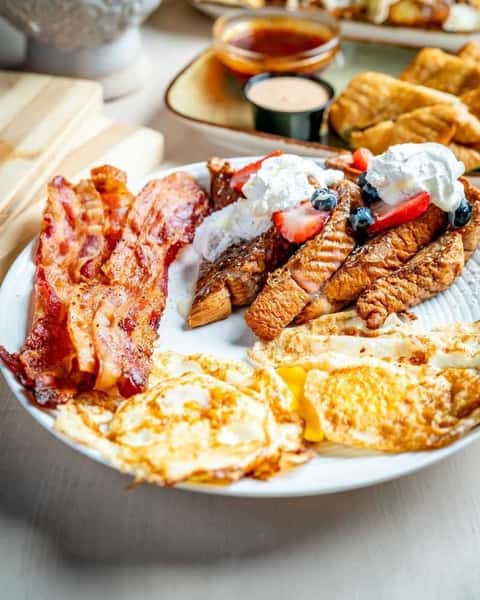 Hearty brunch plate with crispy bacon, fried eggs, and golden French toast topped with whipped cream and fresh berries in a cozy café setting.