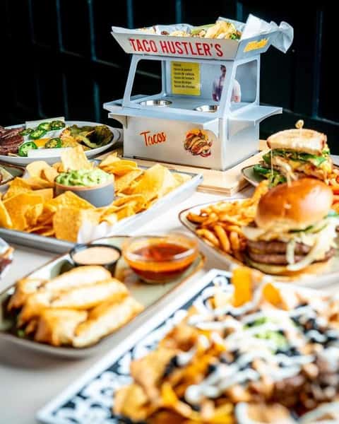 Vibrant restaurant table spread featuring loaded nachos with cheese, black beans, and creamy drizzle, alongside gourmet burgers, fries, and assorted shareable plates in a lively casual dining setting.