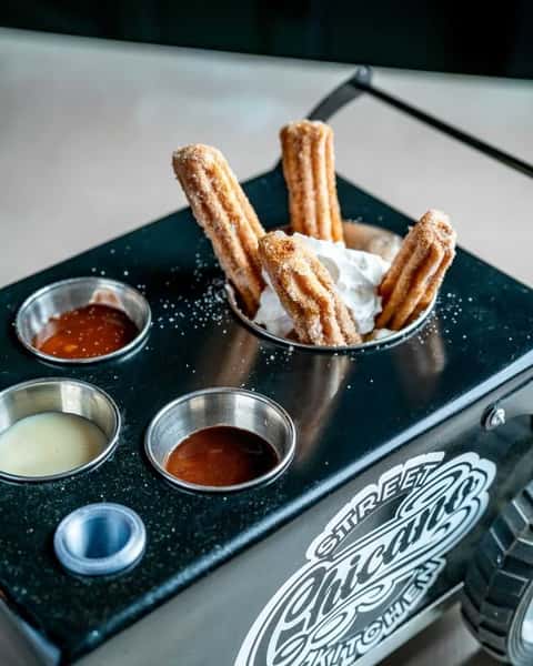Cinnamon sugar churros served with assorted dipping sauces and whipped cream on a street-food style tray, highlighting a fun dessert presentation.