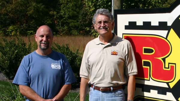 Two men stand next to a sign that says "R & R Grill and Bar-B-Que," which is also on their shirts. The men are smiling, likely owners or managers of the restaurant.