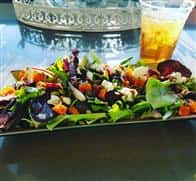 A long plate filled with salad that has a variety of toppings. In the background there is an ice tea with a straw in it.