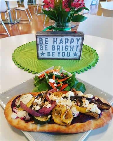 Naan bread with grilled vegetables on top of it paired with a salad next to a sign that says "Be happy, Be bright, Be you".