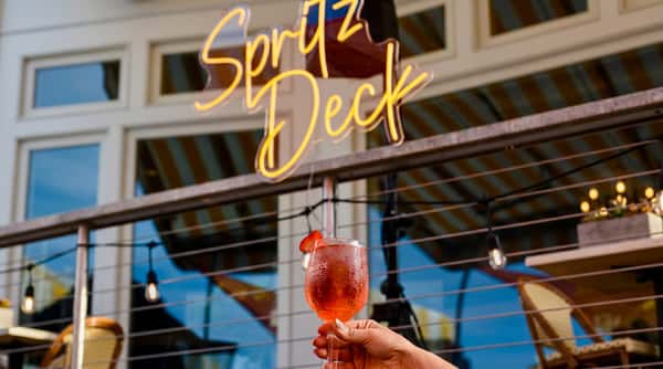 Hand holding an Aperol Spritz with a strawberry garnish under a glowing yellow "Spritz Deck" neon sign.