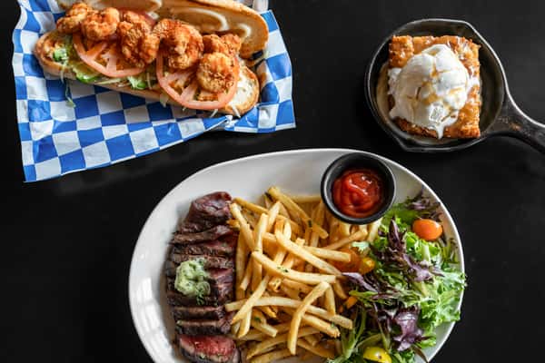 Southern-inspired fried shrimp po' boy sandwich with lettuce and tomato on a hoagie roll, served alongside a steak frites platter with seasoned fries, mixed greens, plus a warm skillet dessert topped with vanilla ice cream and caramel drizzle.