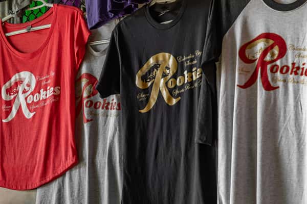 Display of Rookies Sports Bar and Grill branded t-shirts in various styles and colors, featuring the signature logo and 'Columbia City, Seattle' text, hanging on a merchandise rack.