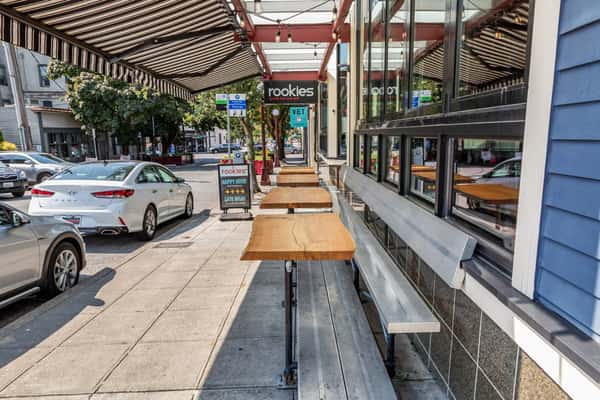 Sidewalk view of Rookies Sports Bar and Grill featuring outdoor bench seating with high-top tables under striped awnings, located in a walkable neighborhood with nearby parking.