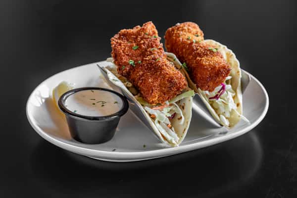Two crispy fried fish tacos filled with shredded cabbage in flour tortillas, served with a side of creamy ranch dipping sauce and a lemon wedge on a white plate.