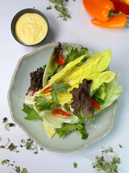 A fresh salad featuring romaine and mixed greens, cherry tomatoes, and a small bowl of creamy dressing, surrounded by colorful bell peppers and herbs on a light background.