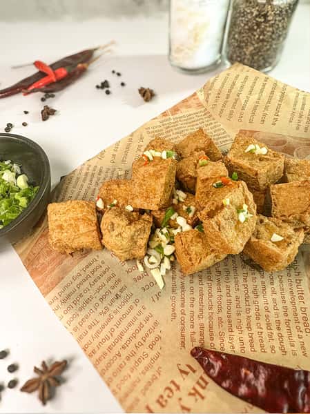 A serving of golden fried tofu cubes garnished with chopped green onions and red chili, placed on a sheet of newspaper, with a bowl of chopped green onions and dried spices in the background.