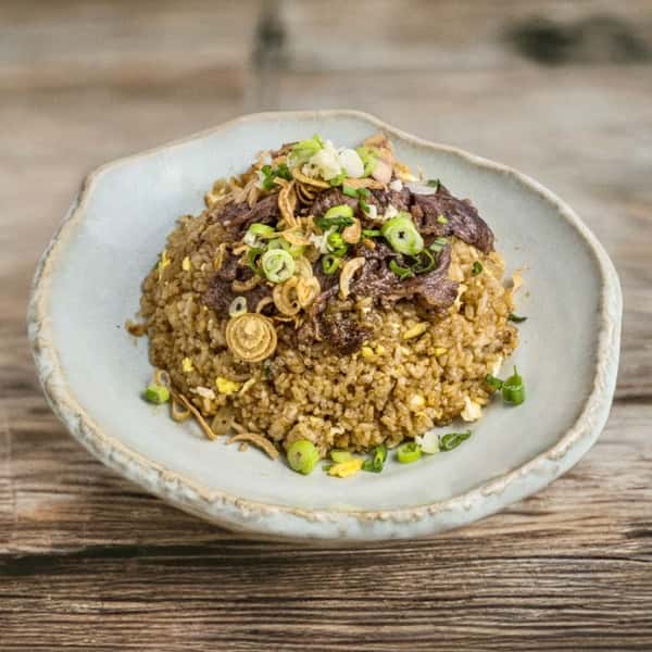 A plate of grilled meats including sliced smoked chicken and sausage, garnished with pickles and onions, served over a bed of fried rice, with a small bowl of sauce on the side.
