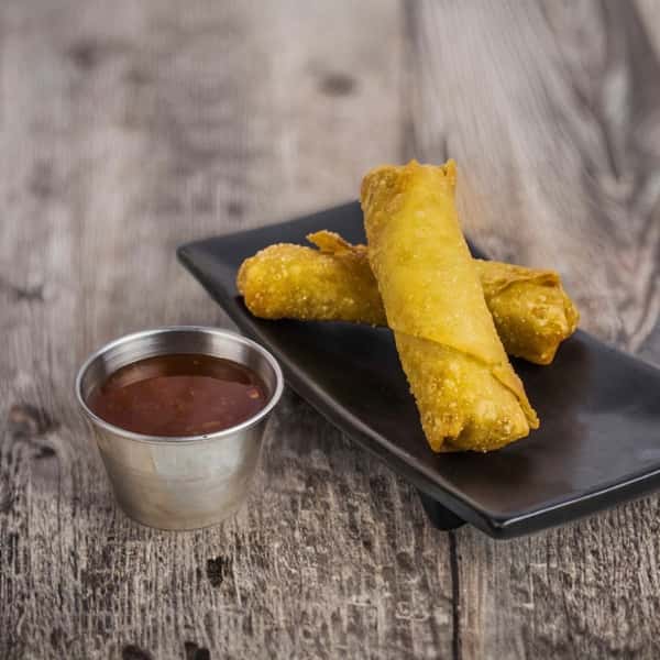 Two crispy spring rolls arranged on a black plate, accompanied by a small metal cup of sweet chili sauce, set against a rustic wooden background