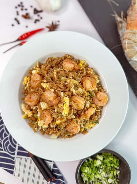 A white bowl filled with shrimp fried rice, featuring pieces of shrimp, scrambled eggs, and green onions, surrounded by red chili peppers and a bowl of chopped green onions on a patterned cloth