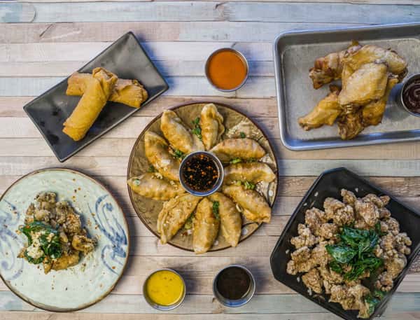 An overhead view of a diverse Asian cuisine spread featuring crispy spring rolls, golden dumplings with dipping sauce, fried chicken wings, and a plate of battered fried mushrooms garnished with greens, all arranged on rustic wooden surfaces.