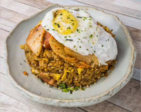 Plate of fried rice topped with a sunny-side-up egg and slices of kimchi, garnished with herbs, served in a light-colored bowl on a wooden table.