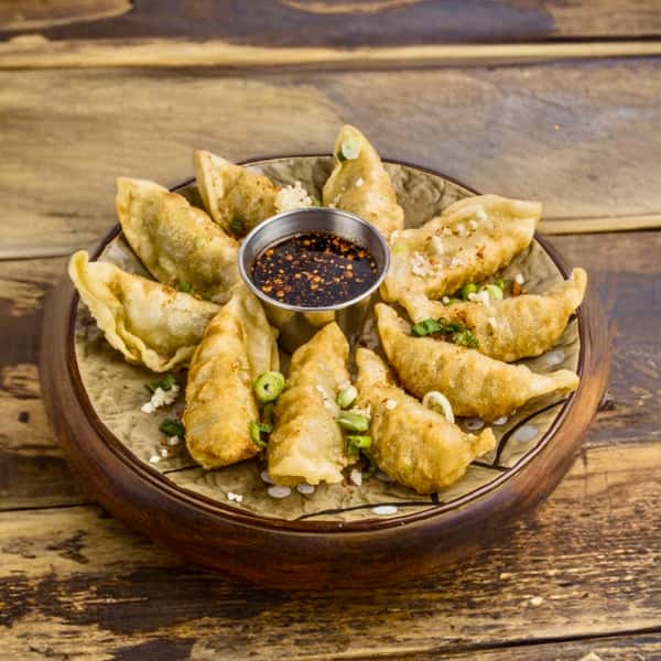 A plate of crispy dumplings garnished with green onions, served with a small bowl of dark dipping sauce, from Pepper Salt in Houston, TX. The plate is presented on a wooden surface, highlighting an inviting and delicious appetizer.
