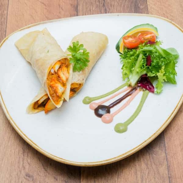 Grilled chicken wrap cut in half and served with a fresh mixed green salad, cherry tomatoes, lemon slices, and colorful sauces on a white plate.