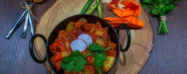Traditional Indian curry with chicken and potatoes, garnished with fresh cilantro and radish slices, served in a black handled bowl on a wooden serving board with fresh vegetables and spices.