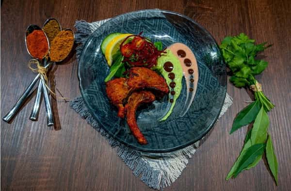 **Alt text:** *Plated tandoori-style lamb chops served with green herb sauce, creamy pink sauce, fresh greens, shredded beetroot, cherry tomatoes, and lemon wedges on a dark blue plate, with spices and fresh herbs arranged beside the dish on a wooden table.*