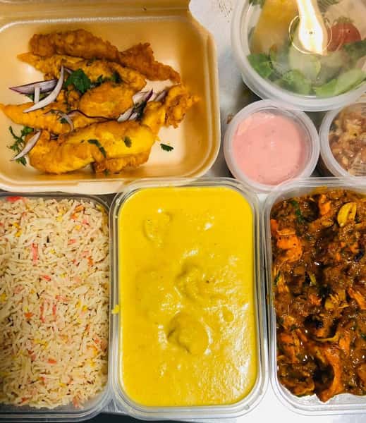 Indian takeout meal with fried chicken, basmati rice, creamy yellow curry, spicy chicken curry, fresh salad, chickpeas, and dipping sauces in takeaway containers.