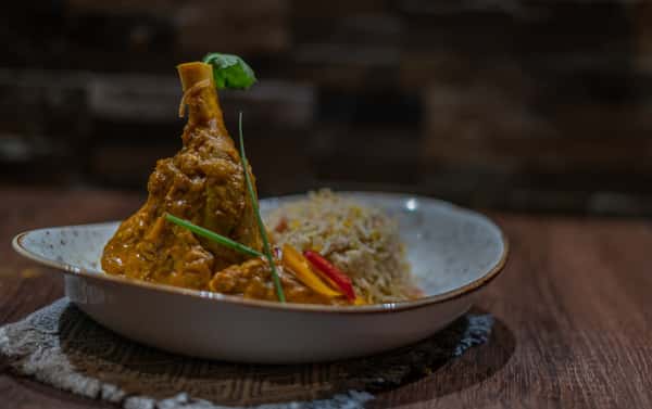 Bone-in chicken curry served with seasoned basmati rice, garnished with fresh cilantro and sliced peppers in a shallow white bowl on a wooden table.