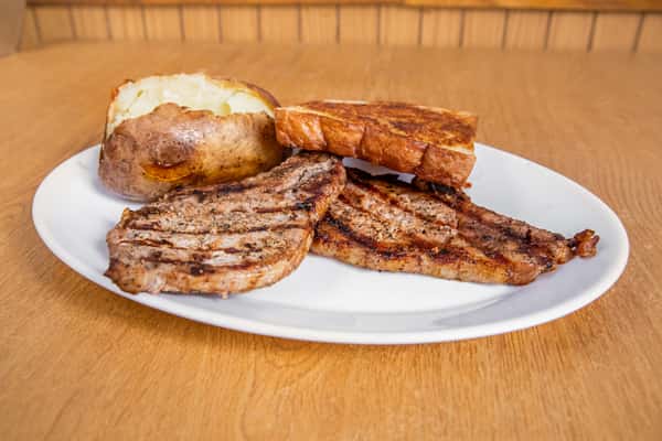 Two grilled pork chops with toast and a baked potato, viewed from the side.