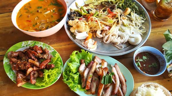 Assorted Thai dishes on a wooden table including tom yum soup, grilled meats, noodles with vegetables, dipping sauce, and fresh herbs.