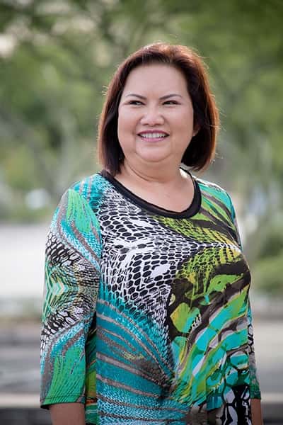 Smiling woman in a colorful green, blue, and black patterned top standing outdoors with trees in the background.